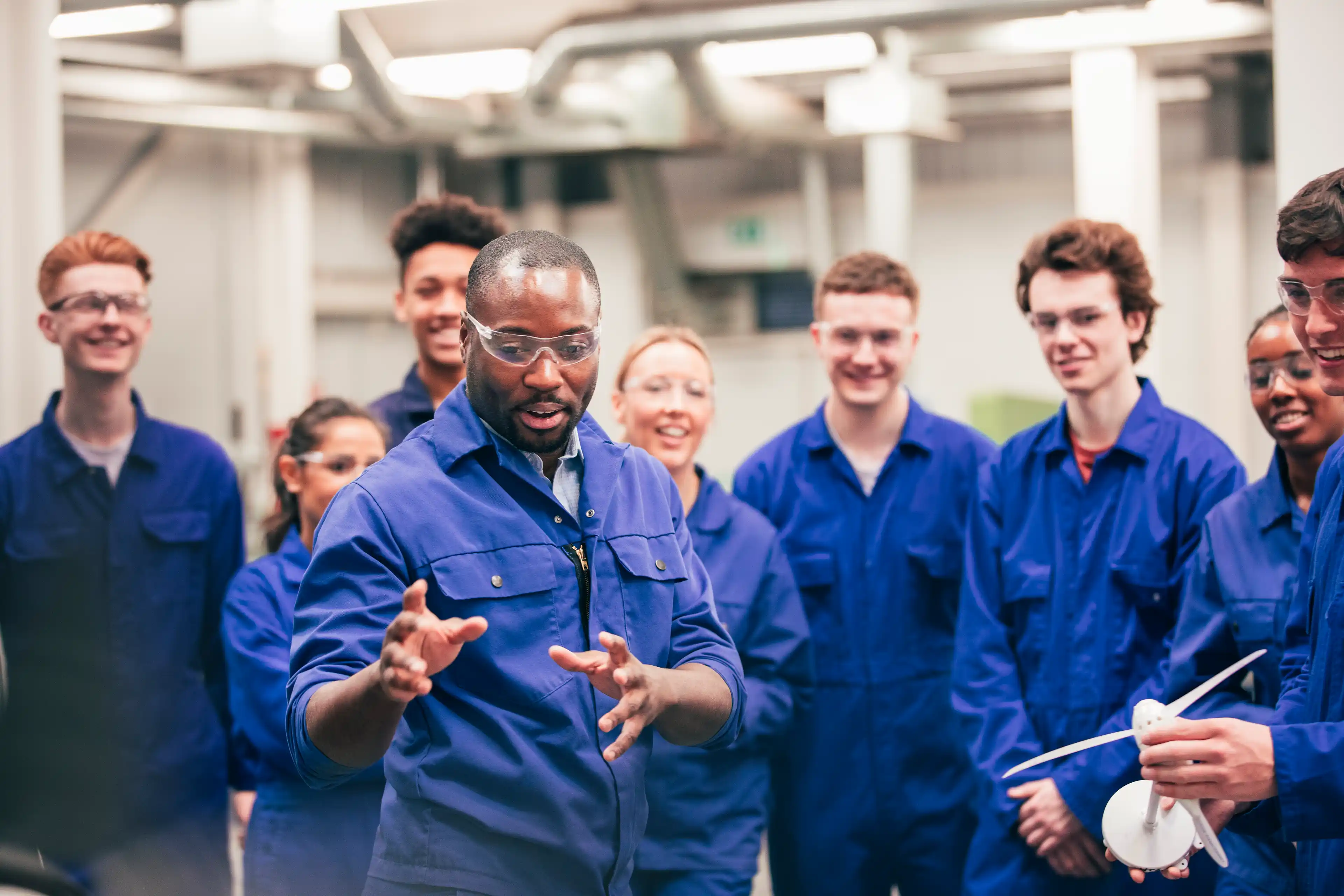 A workshop leader demonstrates something to a group of students, all wearing blue protective overalls