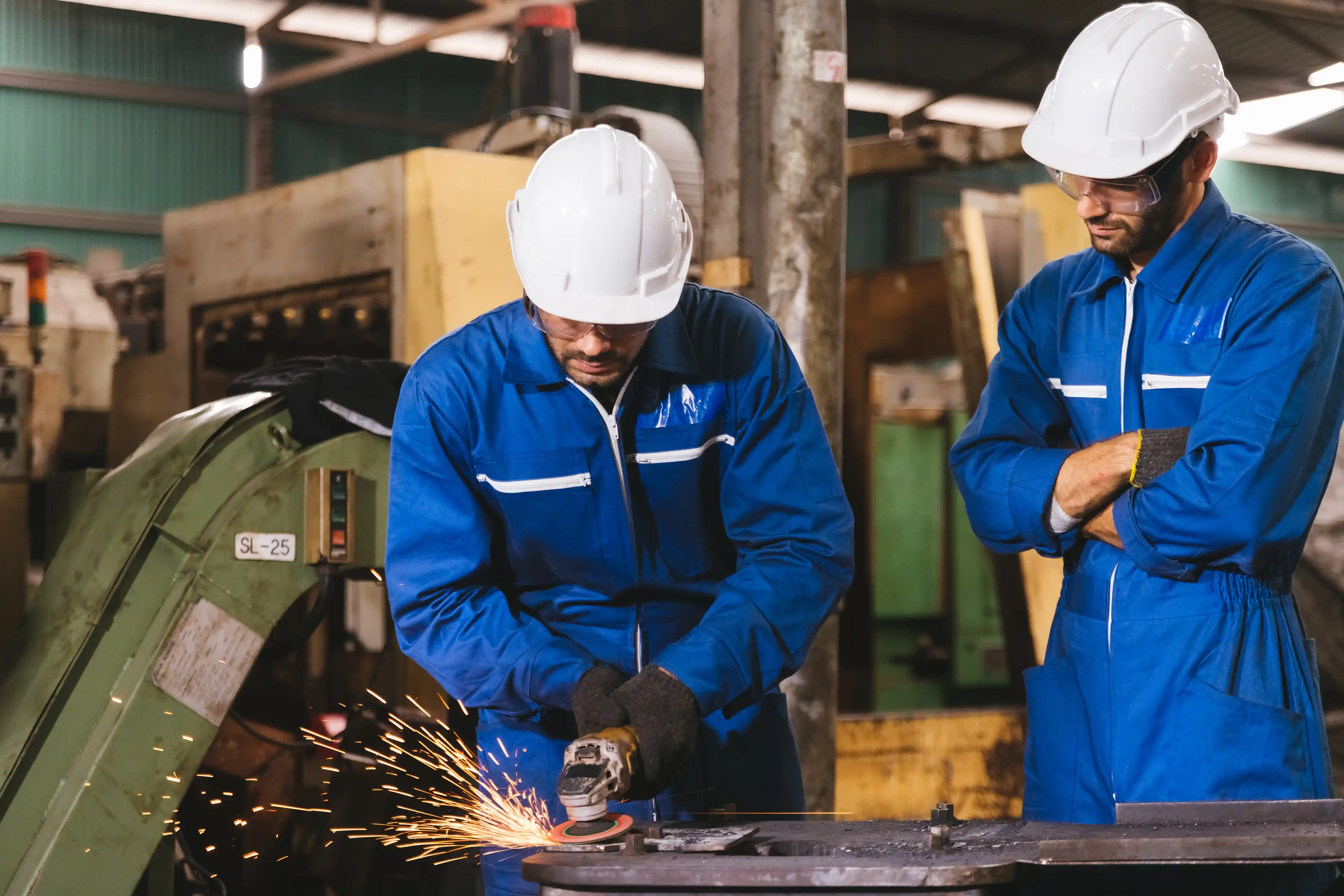 Two technician industrial engineers wearing safety uniform and safety helmet cutting metal part using hand angle grinder machine. Large industrial factory background.