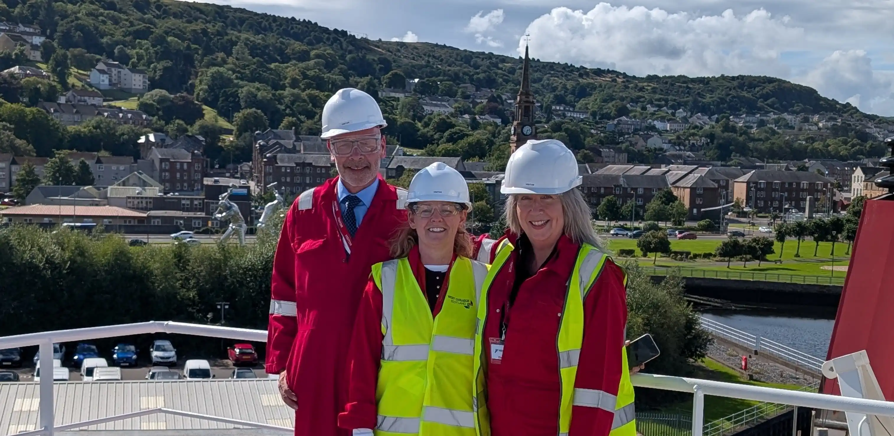 three individuals wearing safety hats, overalls and hi-vis vests with a view of hills behind them