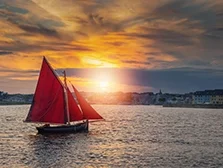 Photograph of a Galway hooker boat with red sail going into Galway harbour at sunset. 