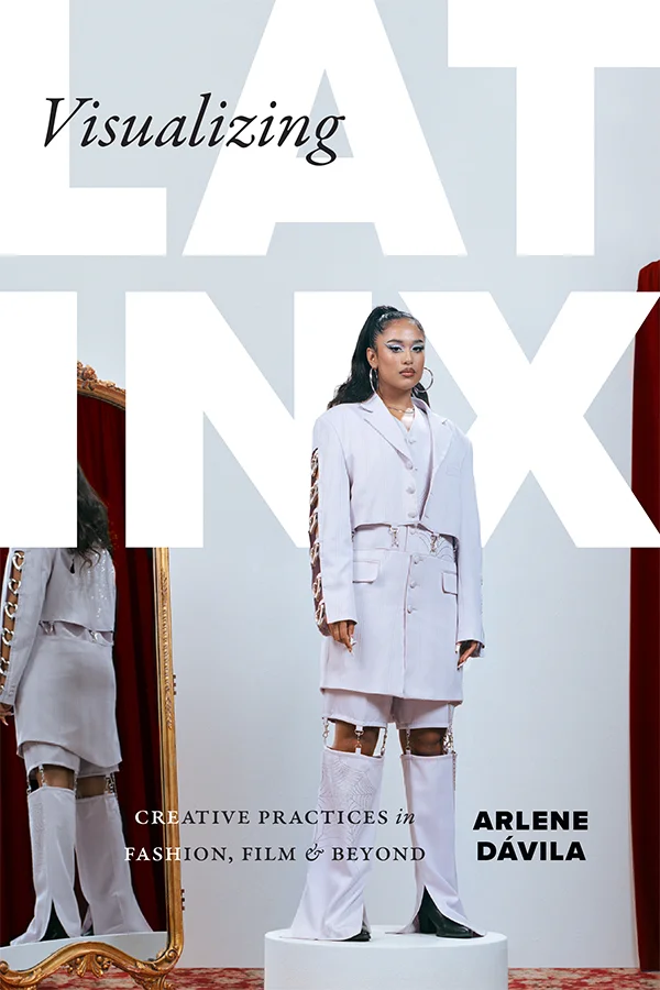 Cover of Visualizing Latinx shows a model wearing a light grey deconstructed suit. She is standing on a podium with a full-length mirror behind her.