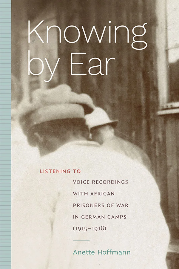 Cover of Knowing by Ear: Listening to Voice Recordings with African Prisoners of War in German Camps (1915–1918) by Anette Hoffmann. Cover features faded black and white imagine of the back of an African American prisoner hunched in front of a wooden building. Title in white text in top left of picture, and subtitle center bottom of picture in red and purple text. Author name below in blue text and similar blue color used as stripped border on the left of the cover.