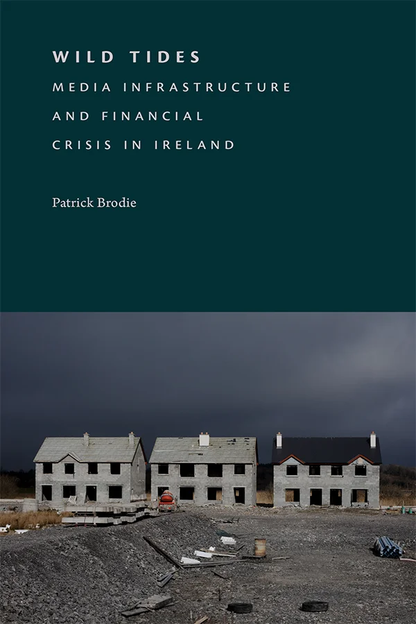 At the bottom of the cover of Wild Tides there is a photograph of three empty houses in disrepair with abandon building materials and gravel in the foreground. The sky is dark and stormy. Above this, the cover is a solid dark teal green.