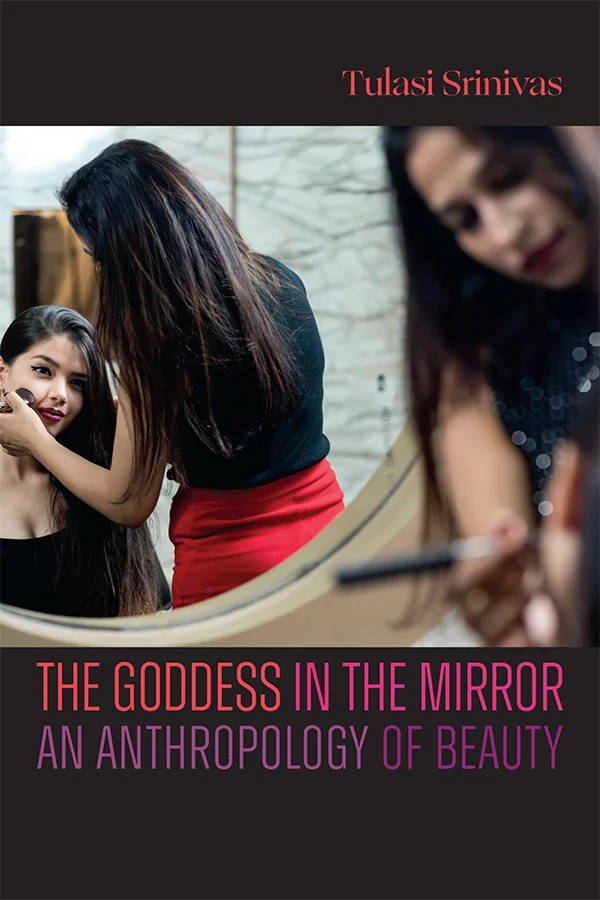 Cover of The Goddess in the Mirror depicts a woman applying makeup to another woman in front of a large round mirror. The scene captures both the women directly and their reflections.