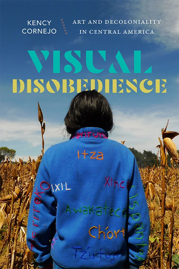 Cover of Visual Disobedience: Art and Decoloniality in Central America by Kency Cornejo. Cover features a photograph of an individual with long black hair looking out over a yellow cornfield. The person wears a blue jacket with various words stitched into it in vibrant colors. The sky is blue above the field with a few scattered groups of puffy, white clouds.