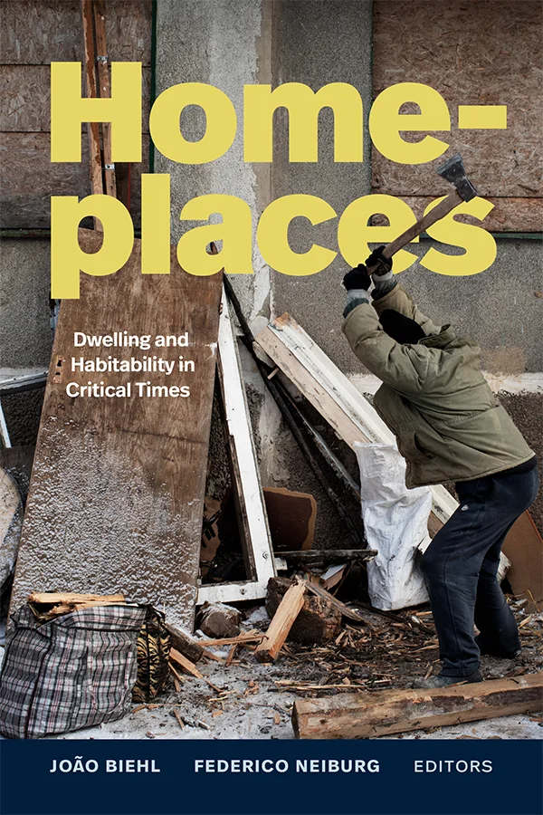 Cover of Homeplaces shows a person dressed warmly swinging an axe overhead while in the process of chopping wood. There is a building with boarded up windows and wooden housing debris laying around.