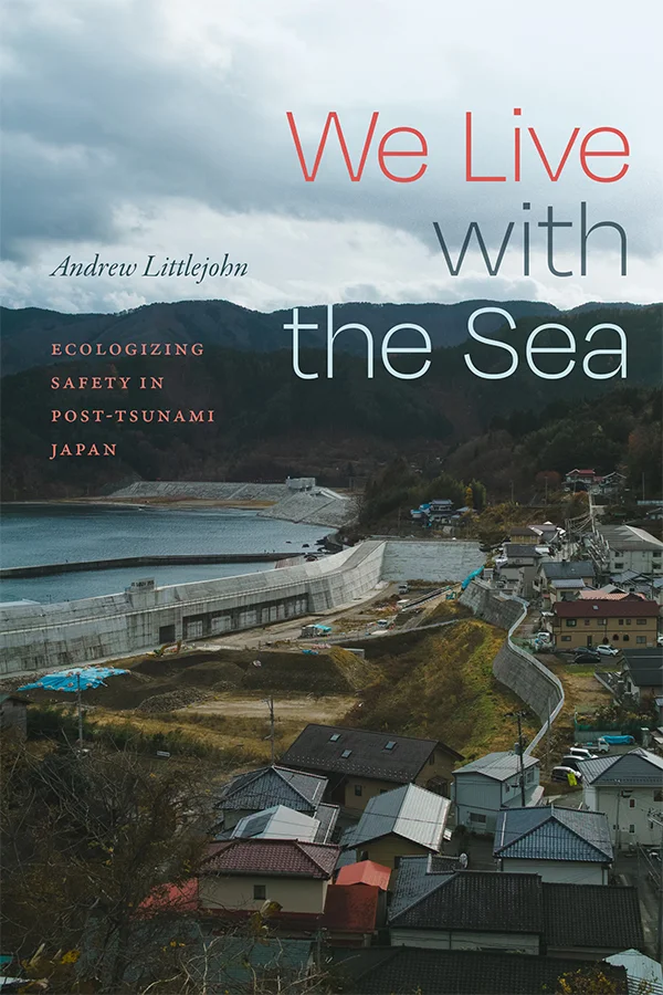 Cover of We Live with the Sea features a photograph of a town in the mountains next to the sea. There are tall concrete walls that are sloped on the sea facing side and separate the town from the water.