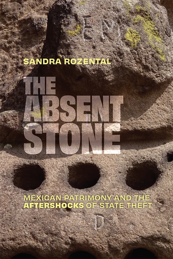 Cover of The Absent Stone features a photograph of part of the Piedra de los Tecomates, a large stone monument. The photo shows three holes in the stone in a horizontal line as well as the letters 'EM' and 'D' carved into the stone.