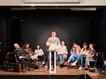 A man in a sweater lectures from a podium while others sit around him looking bored.