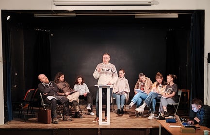 A man in a sweater lectures from a podium while others sit around him looking bored.