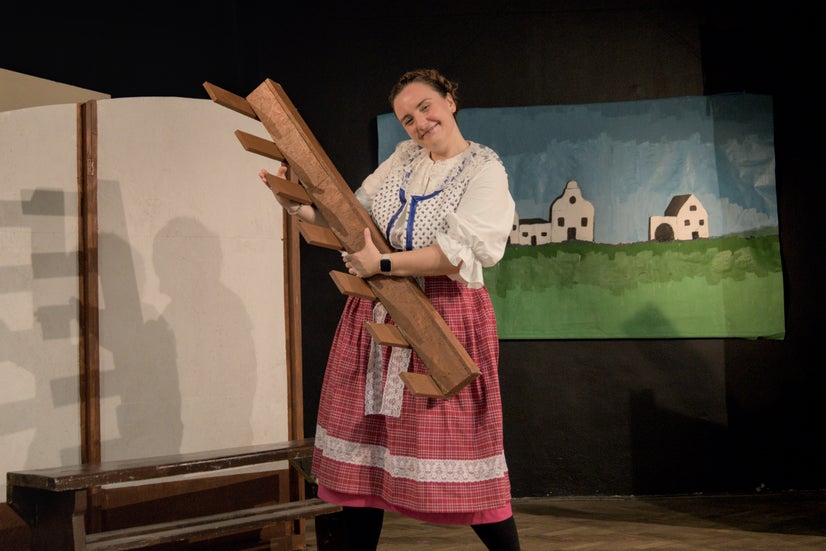 A woman in an old-fashioned white shirt with blue trim and a long, light red skirt smiling and holding a small fence in the air.