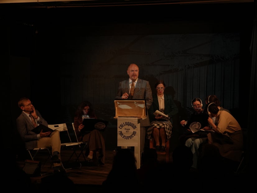 A man gives a speech while on one side another man and a woman think about it. One the other side, a woman in glasses looks annoyed and three women in headsets talk confusedly among themselves.