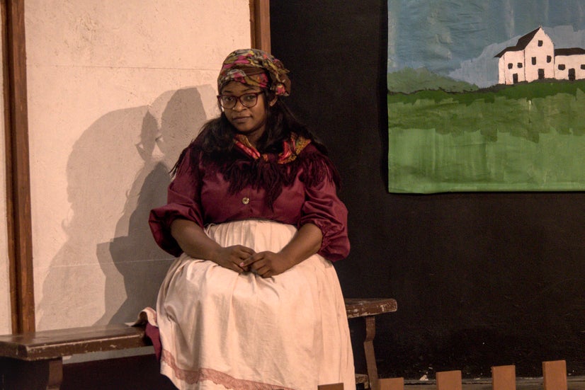 A woman with a dark purple shirt, a scarf with flowers covering her head, and white apron sitting on a bench.