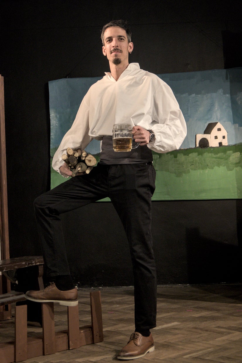 A man in an old-fashioned white shirt standing with one foot one a small fence with a beer in his left hand and some wood in his right.