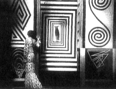 A black-and-white still from an old film, with a woman standing with her back to the camera and geometric shapes painted on the wall.