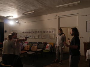 A man points a gun at another man while a woman whispers in his ear. Another woman looks scared nearby.