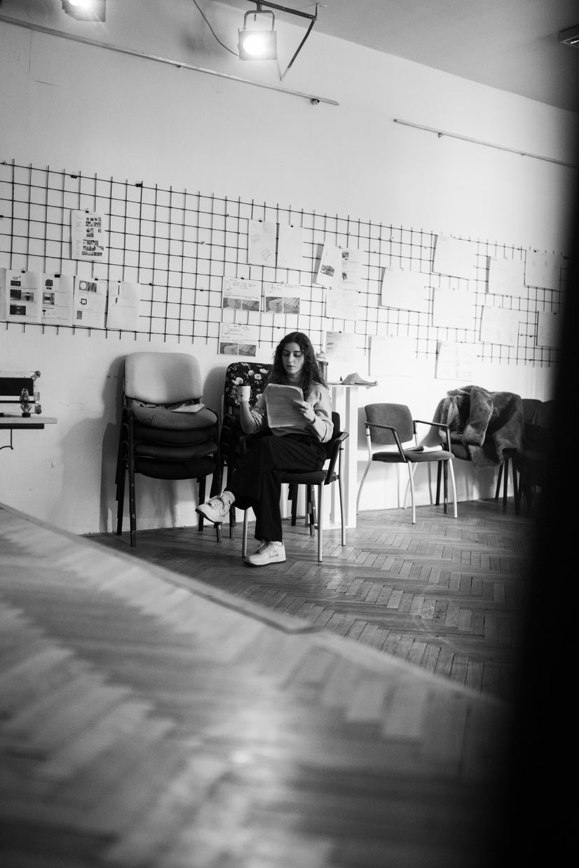 A woman sits in a chair with a script in one hand and a coffee in the other. She seems small with no one else in the picture.