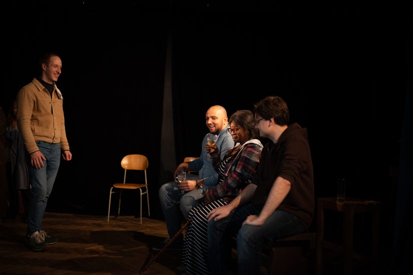 A man stands on the left while three people sit on a bench on the right. The woman in the middle of the bench is raising a glass in a toast and everyone is smiling.