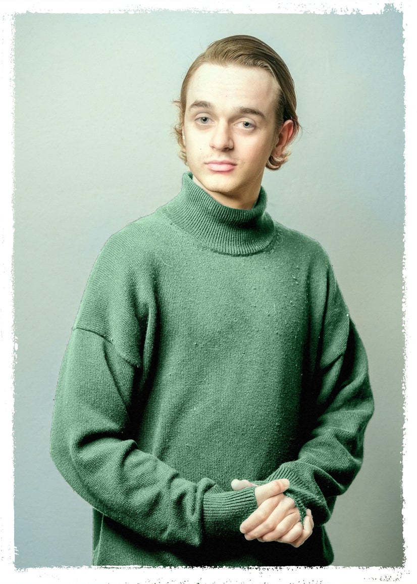 A man in a green turtleneck sweater hands his hands in front of himself and looks at the camera.