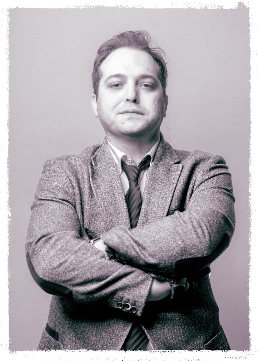 A black-and-white photo of a man in a suit and tie crossing his arms and leaning away from the camera.
