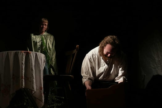 A women in green looks on sadly from the background at a man in white kneeling and crying.