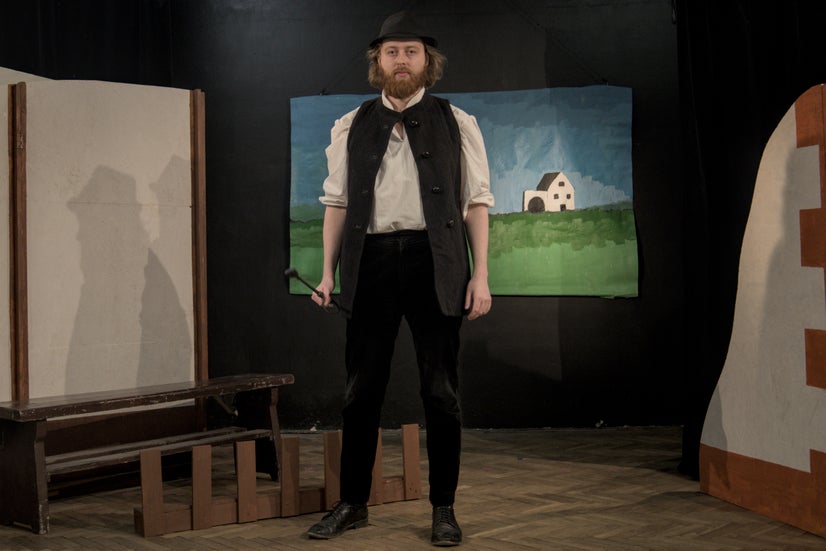 A man in an old-fashioned white shirt with a dark vest and black hat staring menacingly at the camera while holding a whip in his right hand.