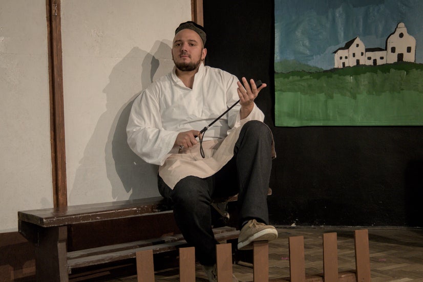 A man in an old-fashioned white shirt with a black cap sitting on a bench behind a small fence smacking a whip against his palm.