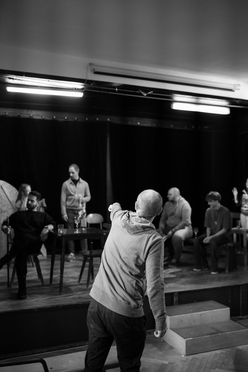 A man facing the stage points to the something on stage, while many actors sit and stand on the stage.