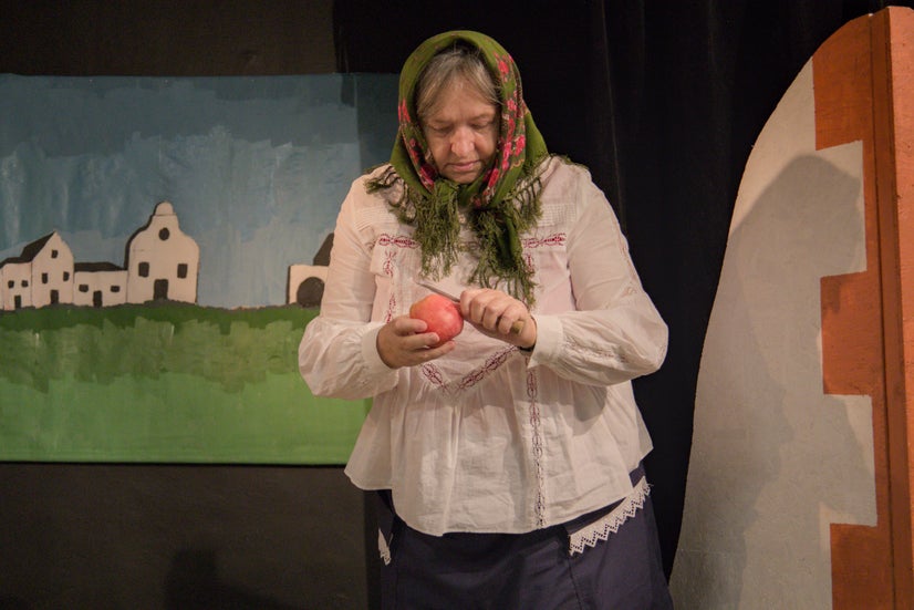 An old woman in an old-fashioned white shirt and green scarf covering her head standing and peeling an apple with a knife.