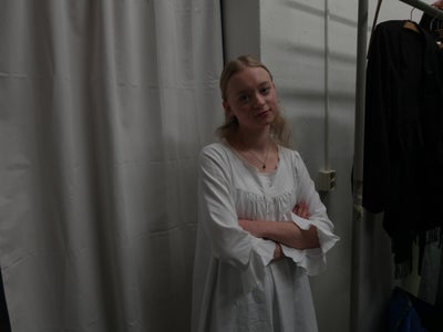 A woman in a flowy white dress stands with her arms crossed in front of a white curtain.