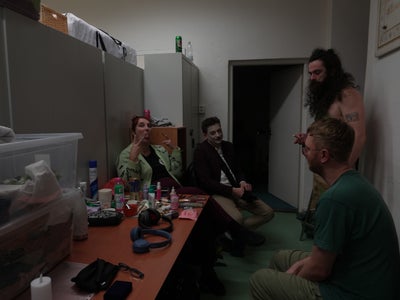 Actors sit in costumes backstage, while the make-up artist sticks out her tongue.