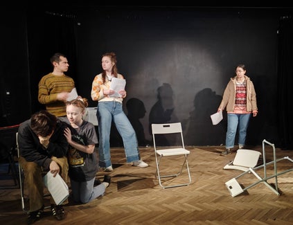 A girl comforts someone crying while others stand at the back of the stage and chairs lie overturned on the stage.
