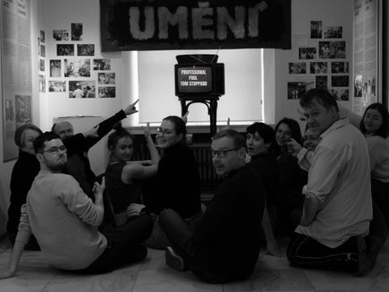 A group of people sit on the floor pointing at an old TV with the words "Professional Foul by Tom Stoppard".