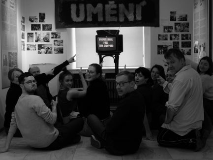 A group of people sit on the floor pointing at an old TV with the words "Professional Foul by Tom Stoppard".