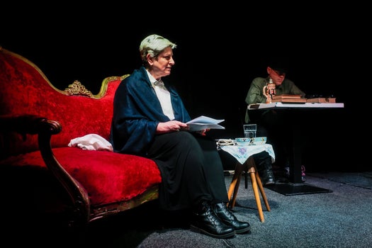 A woman sitting on a red couch in light with a man in a black hat in shadows at a table behind her.