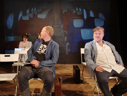 Two men sit in chairs, with one looking over his shoulder at a woman sitting in a chair in the back.