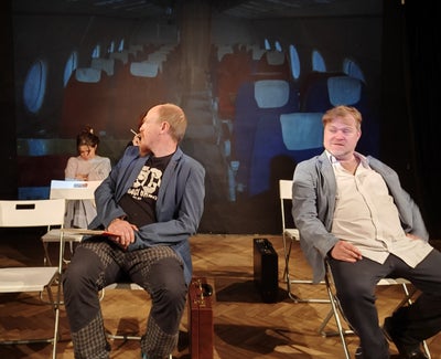 Two men sit in chairs, with one looking over his shoulder at a woman sitting in a chair in the back.
