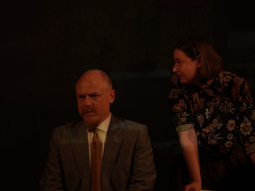 A man in a suit looks away from a woman who is standing and leaning over him.
