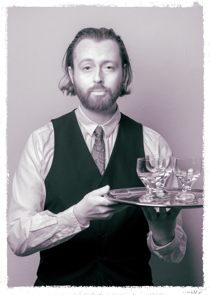 A man in a collared shirt, tie, and vest holding a tray of glasses.
