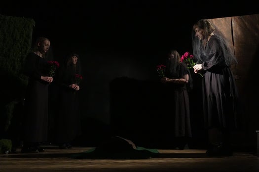 Four women in black with veils gathered in a circle holding red flowers in their hands.