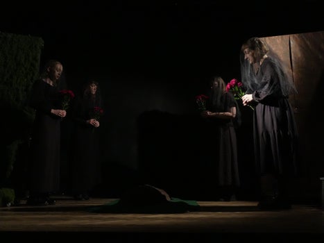 Four women in black with veils gathered in a circle holding red flowers in their hands.