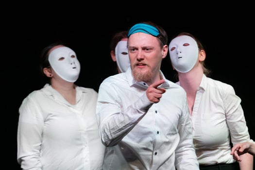 A man in a white shirt with a blue sleeping mask on his head points angrily, while other people in white shirts and white masks stand behind him.