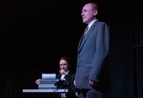 A man stands in the foreground while a woman writes at a typewriter at a desk with a phone.