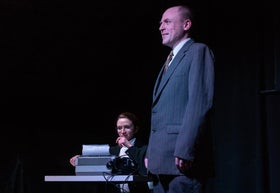 A man stands in the foreground while a woman writes at a typewriter at a desk with a phone.