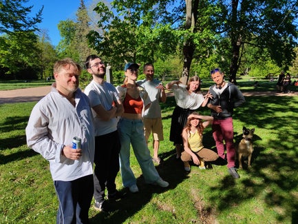 A group of people stand (or squat) in a semi circle on a sunny day in the park.