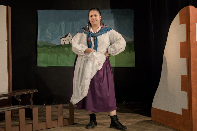 A woman with an old-fashioned white shirt, a blue scarf around her neck, a white apron, and a long purple skirt standing and giving a disapproving look.