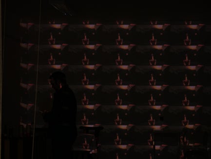 A man appears in shadow in front of a screen with an image repeated many times.