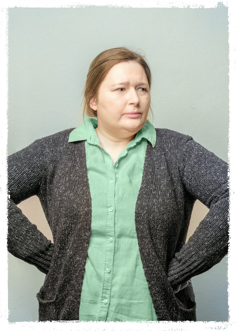 A woman in a sweater with a green shit underneath with her hands on her hips glaring away from the camera.