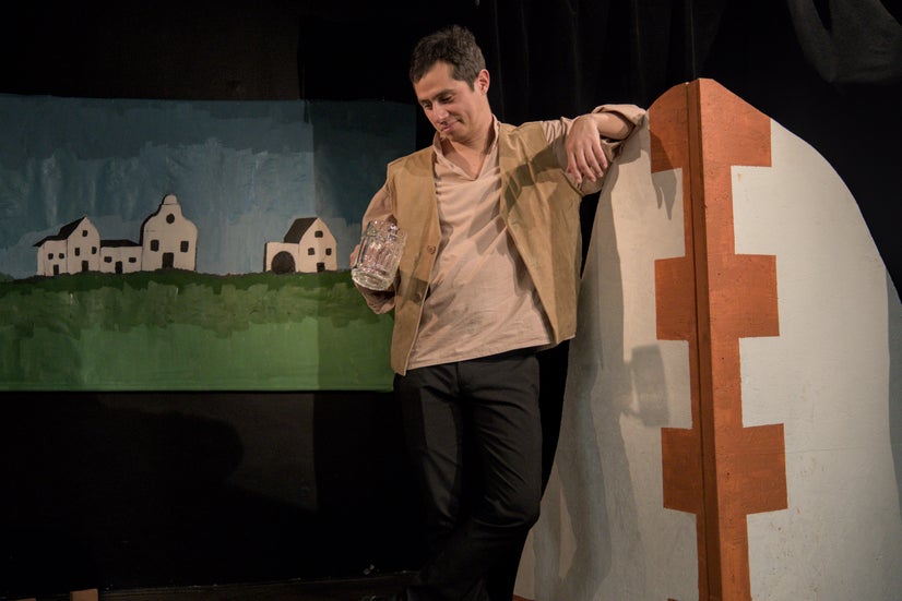 A man in an old-fashioned shirt and brown vest leaning against the end of a stage building looking down at an empty beer mug in his hand with a wry look on his face.