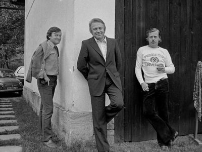 A black-and-white photo of three men. The one on the left is facing a wall with a sports coat around his shoulders. Around the corner, the man in the middle is in a suit with his back against the wall. And on the right is a man in a sweatshirt leaning against a door with a drink in his hand.